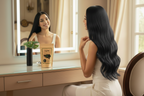 Woman with long hair admiring herself in a mirror with a product box on the vanity.